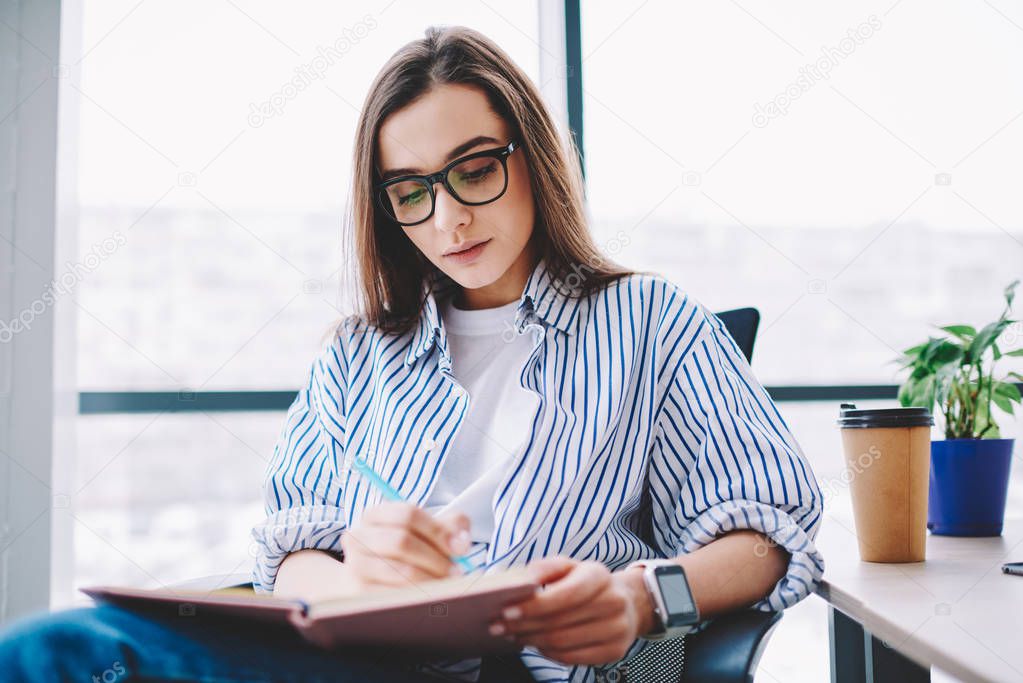 Serious female writer in spectacles concentrated on working process making notes of ideas at desktop, clever brunette woman writing in notepad creating article for publication sitting in offic