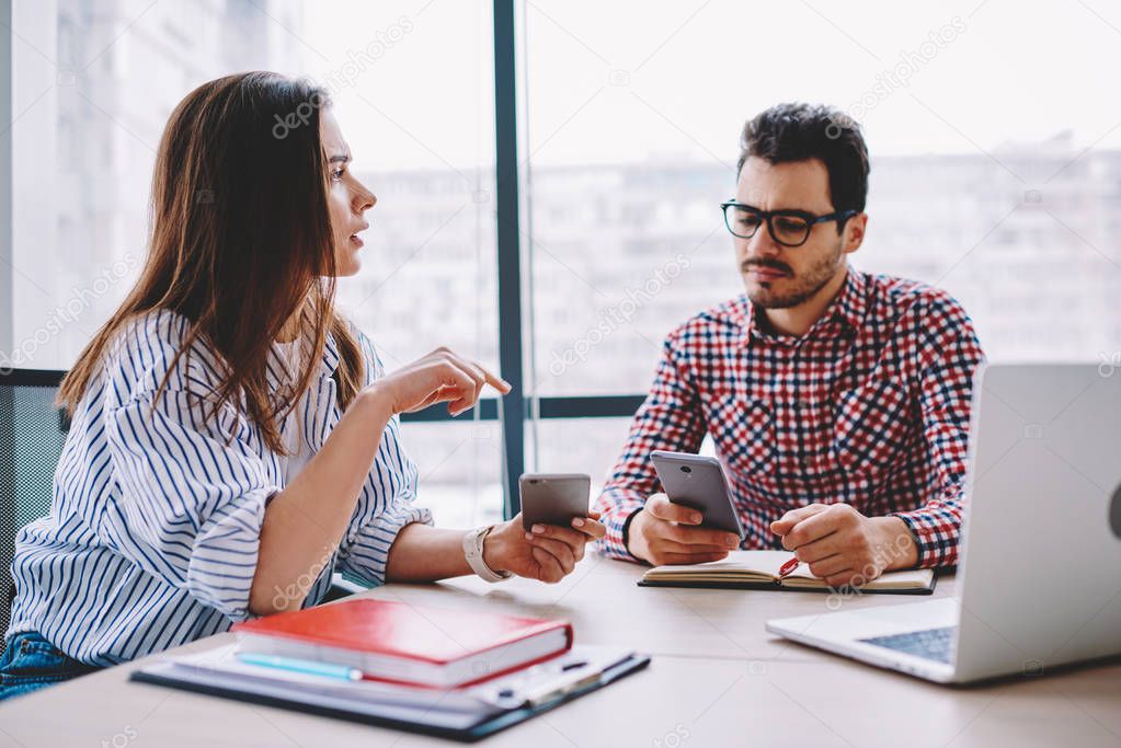 Male and female colleagues talking while share multimedia files via smartphones during working process at desktop, young employees discussing ideas while networking on mobile phones search informatio