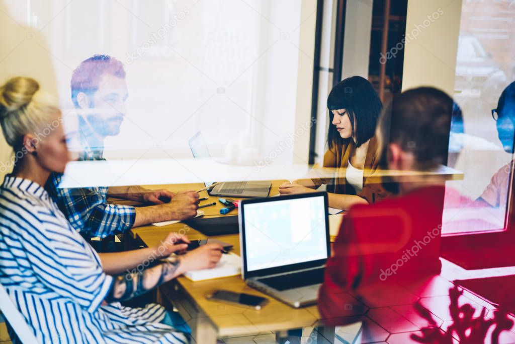 Group of young people in smart casual wear discussing cooperative plan during briefing sitting at meeting table in office behind glass wall.Team of designers collaborating on creative project