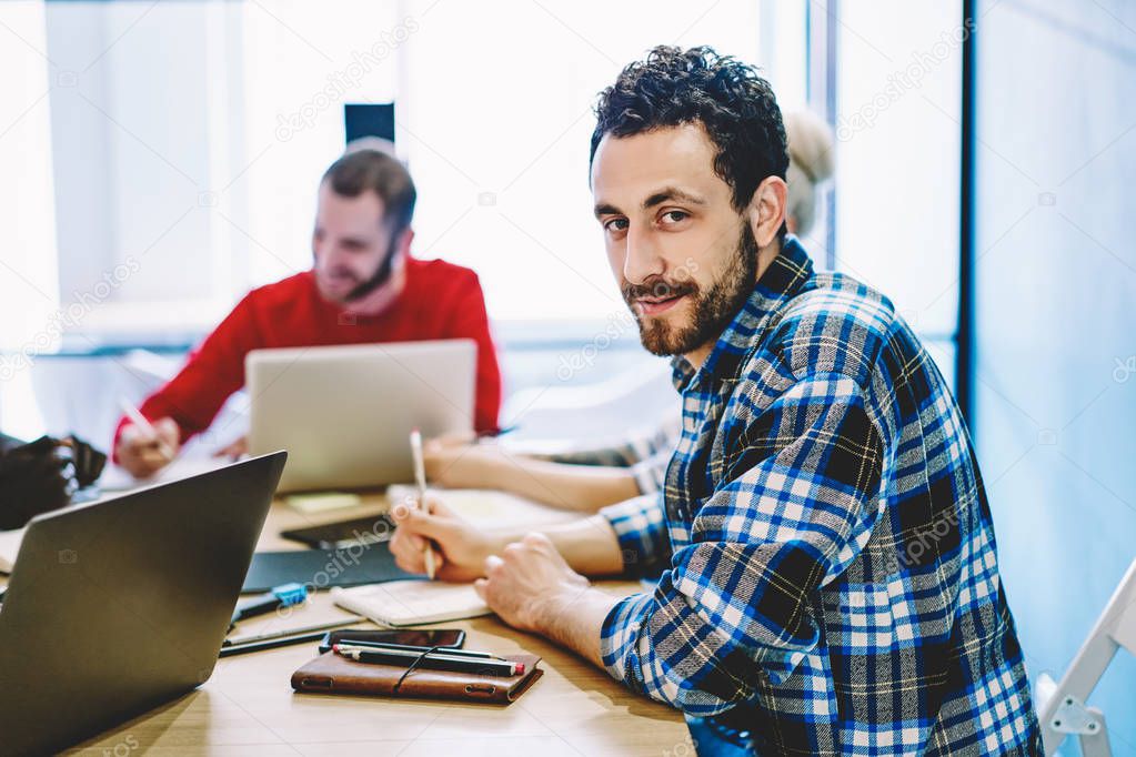 Portrait of young man dressed in casual wear looking at camera while sitting at meeting table and discussing working process in office.Handsome hipster student during studying courses
