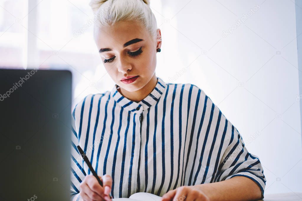 Concentrated young woman dressed in stylish casual apparel writing down information and doing homework in coworking space.Pensive student making notes preparing for studying seminar in college