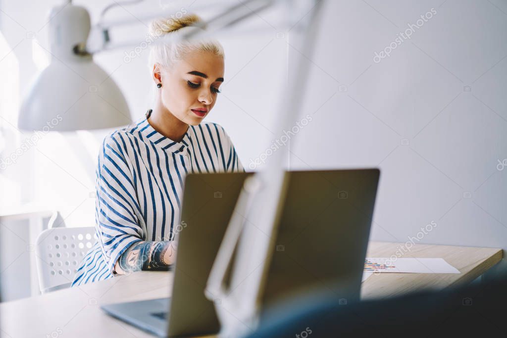 Stylish young blonde woman working remotely in modern coworking space.Casual dressed caucasian hipster girl sitting at desktop with digital laptop computer connected to wireless 4G internet in office