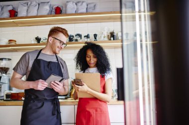 Cheerful young man together with african american hipster girl checking offer on smartphone in modern coffee shop.Successful male and female barista dressed in red apron using 4G internet on phone