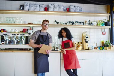 Profesyonel erkek ve dişi barista şık önlük ayakta modern kahve makinesi arka plan üzerinde kendi kafeteryasında tam uzunlukta portresi. Çok kültürlü garson ve kameraya gülümseyen garson
