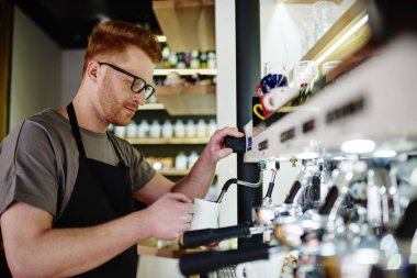 Kendi kahve dükkanında çalışan ve iş başlangıç gelişen modern makinede lezzetli kahve içecek hazırlama önlük beyaz erkek profesyonel barista. Dalgın genç adam garson kapuçino yapma
