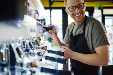 Pozitif beyaz erkek barista lezzetli içecek modern kahve makinesi üzerinde kendi kafeteryada yaparken gülüyor. Kafede içki hazırlarken eğleniyor neşeli deneyimli garson