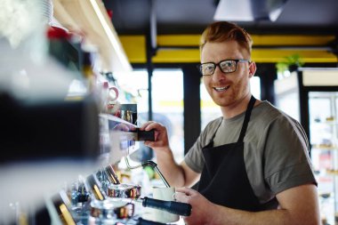 Modern makine ayakta çalışma yerinde kullanarak içecek hazırlarken kameraya gülümseyen önlük profesyonel barista portresi kendi kafeterya bar. Pozitif genç adam garson kahve içecek yapma