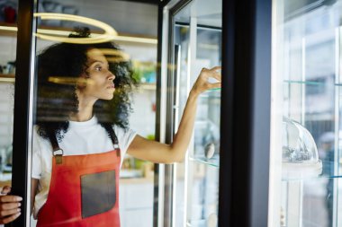 Afrikalı-Amerikalı kadın pudra buzdolabı kendi fırın satmak için kek ile kontrol. Koyu tenli Genç kadın kırmızı önlük şeker çubuğu ayakta şık kafeteryada açılış yoğunlaştı