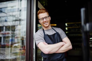 Kızıl saçlı ve siyah önlük kameraya gülümseyen başarılı ve profesyonel erkek barista portresi. Çapraz elle kendi kahve dükkanı için anterence adlı dururken gülüyor olumlu beyaz garson