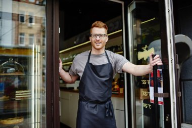Önlük çalışan garson ve kendi kahve dükkanı vitrin üzerinde tatlı kek ile açılış kapı olumlu beyaz sahibi. Kendi Cafe ayakta girişinde Kızıl saçlı davet ile neşeli erkek barista