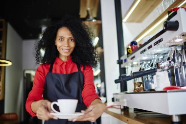 Lezzetli kahve veren ve kameraya gülümseyen Afro-Amerikan olumlu barista yarım uzunluk portre. Neşeli garson önlük fincan çevre makine oen cafe bar, aroma içecek ayakta tutan