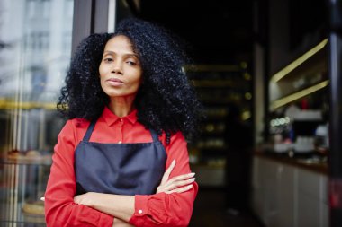 Yarım uzunluk portre profesyonel Afro-Amerikan barista girişte kahve dükkanına ayakta açık havada süre kameraya bakarak kıvırcık saçlı. Kendine güvenen koyu tenli kadın sahibi Cafe