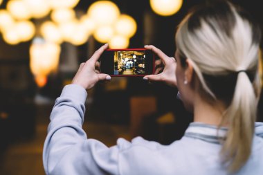 Arkadan görünüşü yatay konum hareket eden telefon içinde tutan ve altın shinning ışıklar yüksek kaliteli dijital fotoğraf makinesinde çekim sarışın beyaz kadın. Genç kadın alarak resim Smartphone cep telefonu ile