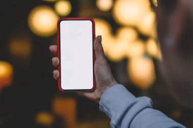Dikiz kadın tutmak içinde dikey konumu cep telefonu bokeh ışıklar akşam zamanında shinning altın karşı elinde. Kadın modern smartphone ile boş mockup kopya alanı perde reklam için tutar