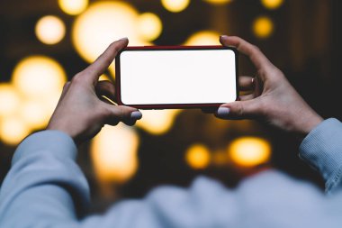 Kadın arkadan görünüm tutun yatay konum cep telefonu altın shinning bokeh ışıklar karşı elinde zaman akşam. Kadın modern smartphone ile boş mockup kopya alanı perde reklam için tutar
