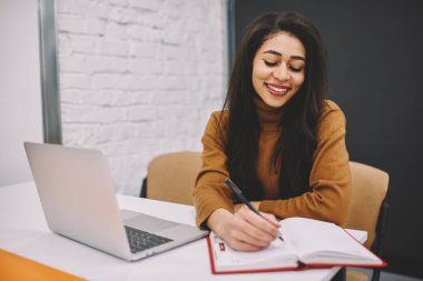 Kız öğrenci dizüstü bilgisayar, onun yayın notları Not Defteri'nde memnun yaratıcı fikirler yapma neşeli kadın blogger yakınındaki masada oturan üniversite sınıf ev ödevleri görevleri yerine getirmeye gülümseyen