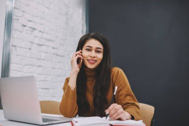 Telefon görüşmesi sırasında cep telefonu görüşmesi sırasında iş bilgilerini belirterek olumlu genç kadın diz üstü bilgisayar ile coworking alanda otururken neşeli kız öğrencinin portresi