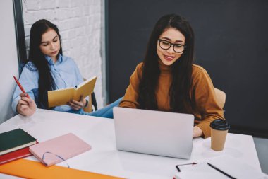 Webinar sırasında onun meslektaşım izlerken ödev öğrenme için dizüstü bilgisayar kullanan akıllı kız öğrenci kitabı, pozitif kadın arkadaş vasıl okul sırası oturan ortak çalışma görev için ile işbirliği