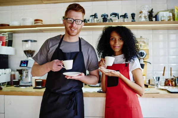 Portrait of two positive male and female colleagues in aprons holding white cups in hands enjoying tasty coffee beverage.Cheerful professional baristas drinking cappuccino during break at bar