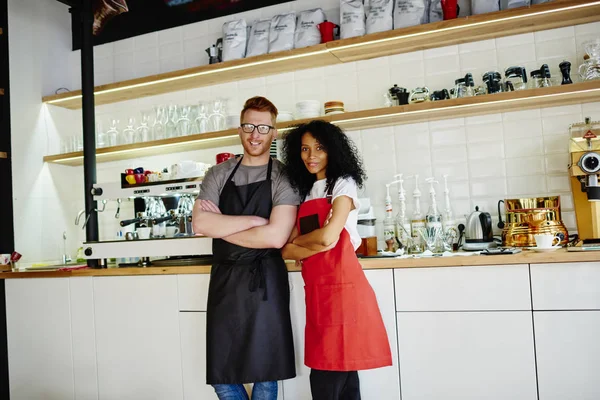 Full length portrait of multicultural team of cheerful barista and waiter with crossed hands smiling at camera.Positive male and female employees in aprons standing at bar and working together in cafe