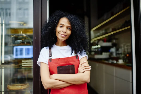 Half length portrait of cheerful african american professional barista in red apron smiling at camera.Positive dark skinned female waitress with crossed hands standing at entrance to own coffee shop