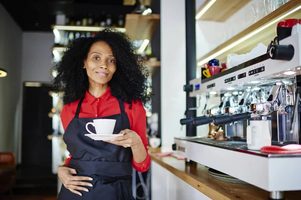 Half length portrait of african american young woman working barista and holding cup of aroma caffeine beverage in hands while smiling at camera standing near modern coffee machine in cafe