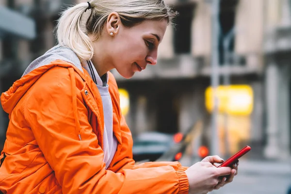 若い女性の街に立っている間携帯電話データ インターネット接続のビューを閉じます スマート フォンでチャット アプリケーション経由でテキスト メッセージを読んでオレンジ ジャケットで白人の内気な少女 — ストック写真
