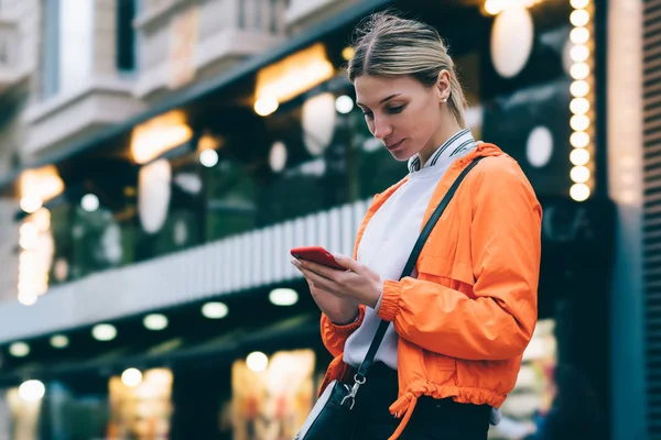 Young female person in bright orange jacket with modern mobile phone device while standing outdoors on city bokeh lights background.Woman typing text message on smartphone using 4G internet connection