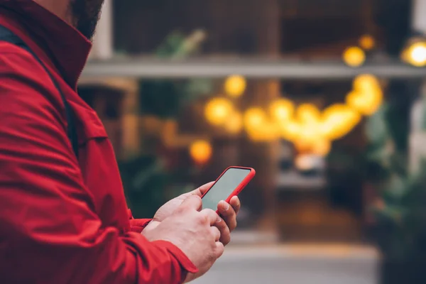 Side cropped view of male person in red jacket touching with finger screen of modern smartphone device outdoors at evening street. Men's hands holding mobile phone near copy space with bokeh lights