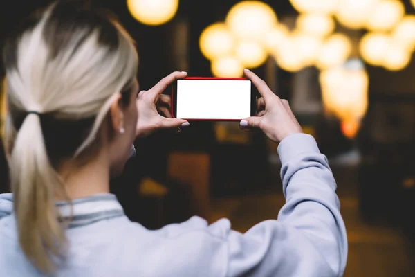 女性手持手在水平位置的背部视图在傍晚时分 手机对金色闪烁的散景灯 年轻妇女保持现代智能手机与空白模拟复制空间屏幕 — 图库照片