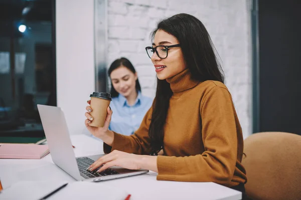 戴眼镜的正黑发女孩在笔记本电脑上工作手持杯咖啡为生产力 微笑的年轻女子在网上观看视频在线上的邮政休息 — 图库照片