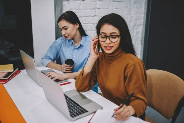 坐在办公室里的年轻妇女集中精力在他们的任务上通过膝上型计算机来拨打电话和监控新闻 忽略了生产具身的沟通 — 图库照片
