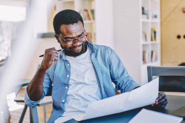 Afro-Amerikan erkek mimar robot resmi planlama, düşünceli akıllı koyu tenli çalışan kontrol bilgileri masaüstünde oturan verileri analiz raporundan şaşkın bakarak konsantre