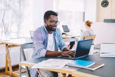 Afro-Amerikan erkek öğrenci dizüstü bilgisayar sosyal ağ kitaplığı'nda wifi kullanarak arkadaş sohbet üzerinde yazarak gülümseyen, netbook ile online iş olumlu koyu tenli freelancer memnun