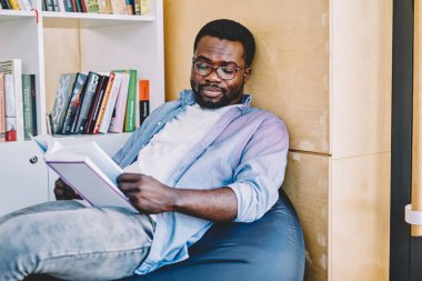 Serious clever african american student in spectacles for vision correction reading interesting book on leisure thinking about plot,dark skinned hipster guy analyzing information from literature clipart