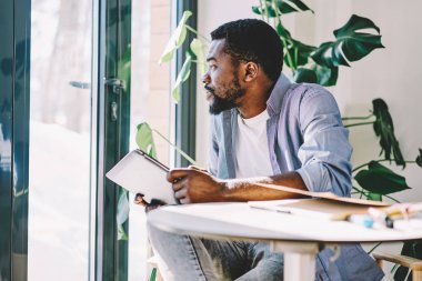 Alan coworking pencere bakarak ağ için dalgın Afro-Amerikan hipster adam holding dijital tablet, ciddi karanlık erkek blogger articl fikirleri hakkında ikram düşünme hakkında rüya derili.