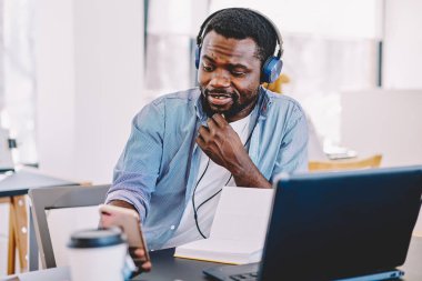 Erkek kulaklık kulaklık Smartphone, Afrikalı-Amerikalı adam 4 g video yapmak için Internet çağrısı hücresel oturuşta üzerinden uzay coworking kullanarak çalma listesinden müzik dalgın koyu derili