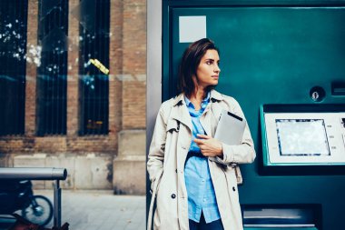 Durulması genç kadın 20 yaşında dijital tablet tutarak ve uzak tramvay durağı kentsel ortamda ayakta beklerken seyir şık ceket giymiş. Hippi kız modern Dokunmatik ped cihaz ile