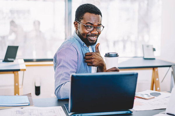 Portrait of cheerful african american male freelancer holding coffee cup showing ok sign enjoy break,successful dark skinned employee at desktop with laptop computer gesture thumb up satisfied with jo