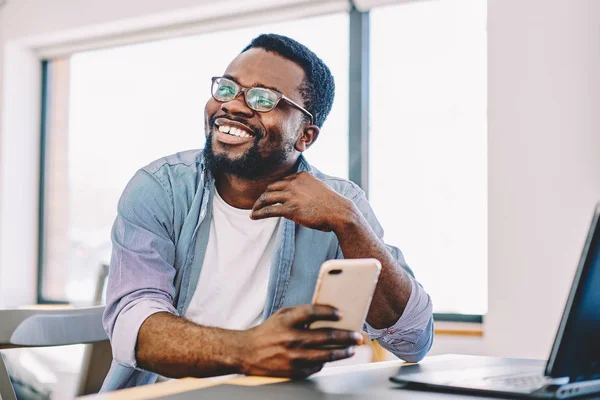 Cheerful african american young male smiling holding mobile phone for checking new feed in social networks, happy dark skinned hipster guy looking away laughing at idea for publication in blo