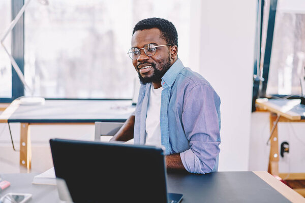 Pensive dark skinned male freelancer in eyewear for vision protection sitting near desktop during remote job, creative african american blogger thinking about idea for update profile in social network