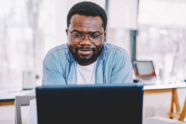 Clever african american male in eyewear for vision protection watching webinar for education on laptop computer,serious dark skinned hipster guy reading information from web page making banking