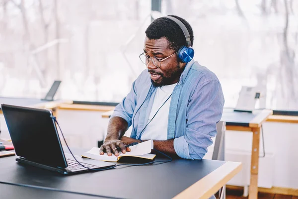 Young african american man in modern headphones listening audio book for education doing homework, dark skinned hipster guy entertaining with playlist on netbook writing ideas for articl
