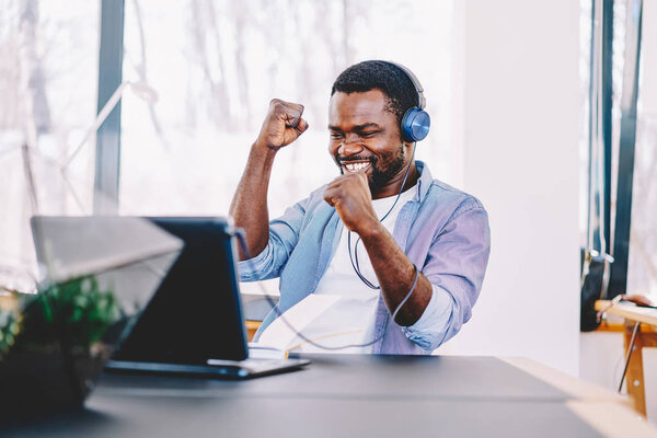 Emotional dark skinned male freelancer celebrating completed project successfully, happy african american football fan cheering for match watching online video on netbook and in headphones in coworking