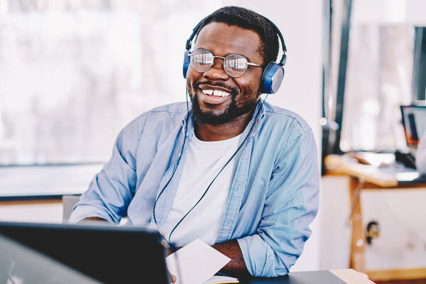 Cheerful african american hipster guy enjoying music in modern headphones and online radio broadcasting on laptop computer, dark skinned male student entertaining with litening playlist on netboo