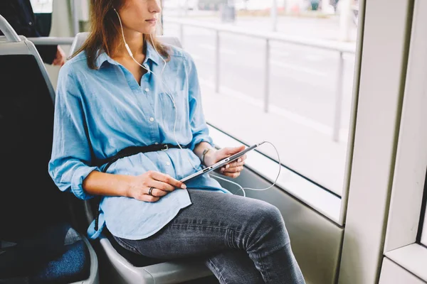 路面電車で座っているモダンなタッチパッドにインストールされた音楽アプリケーションからオーディオの歌を聞くイヤホンで若い女性を熟考のビューをトリミングしました 内気な少女が公共交通機関を使用して無料の無線インターネット — ストック写真