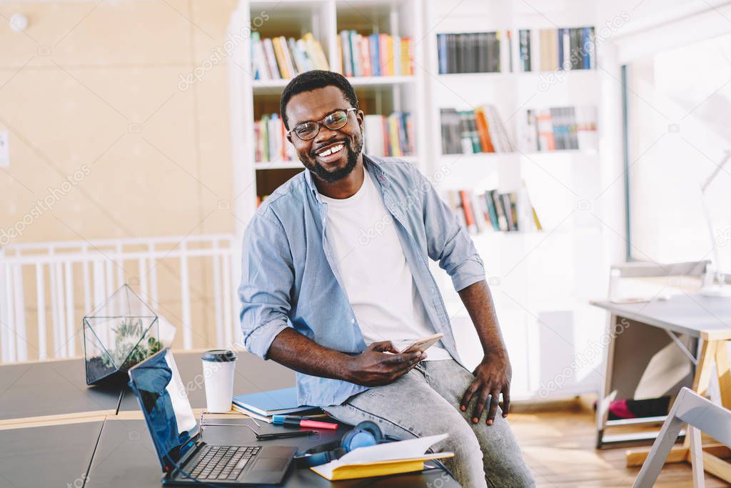 Portrait of cheerful african american male entrepreneur satisfied with success on job sitting on working place using mobile phone,happy prosperous businessman in casual wear looking at camera