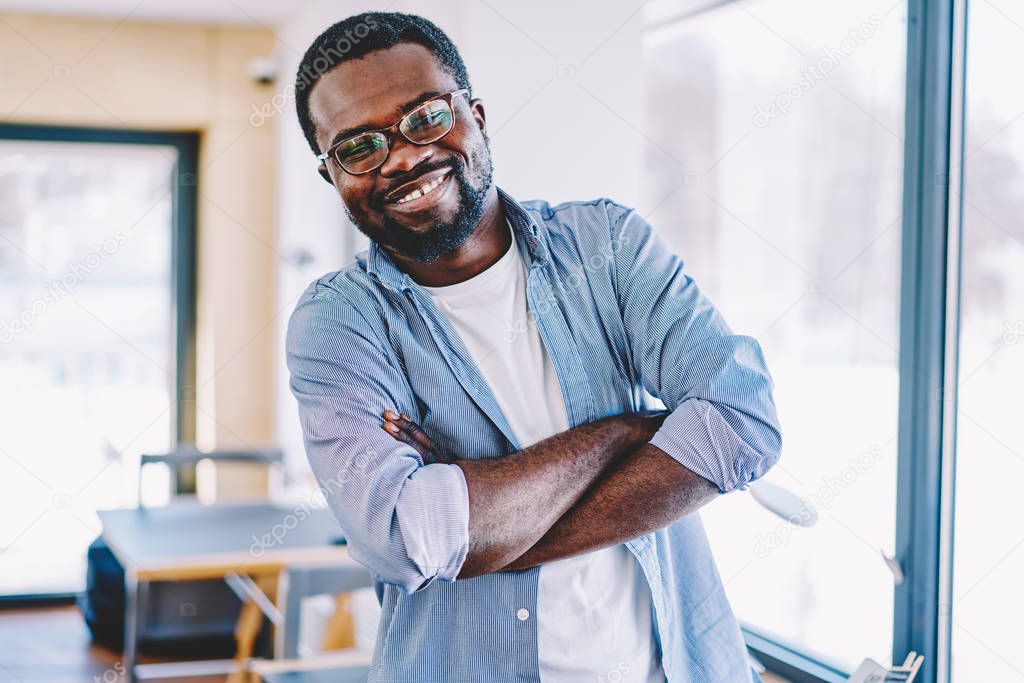 Half length portrait of cheerful dark skinned male entrepreneur in casual wear standing with crossed arms satisfied with completed job,african american funny guy laughing and looking at camer