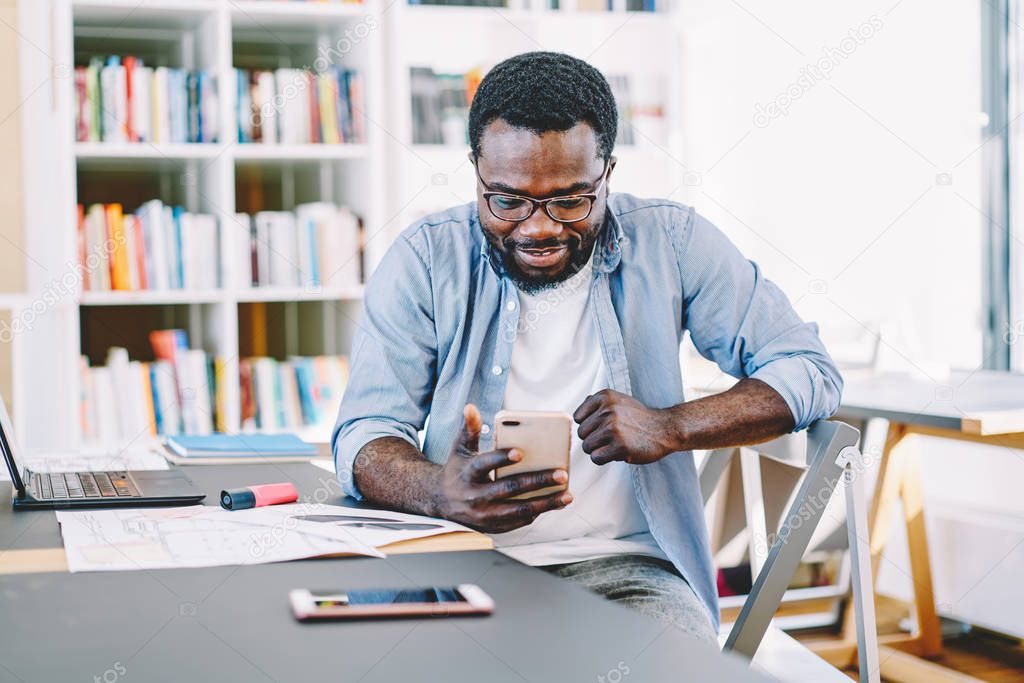 Cheerful african american hipster guy using smartphone for watching funny video  on work break, positive dark skinned male chatting in app on modern cellular checking notification in coworking space
