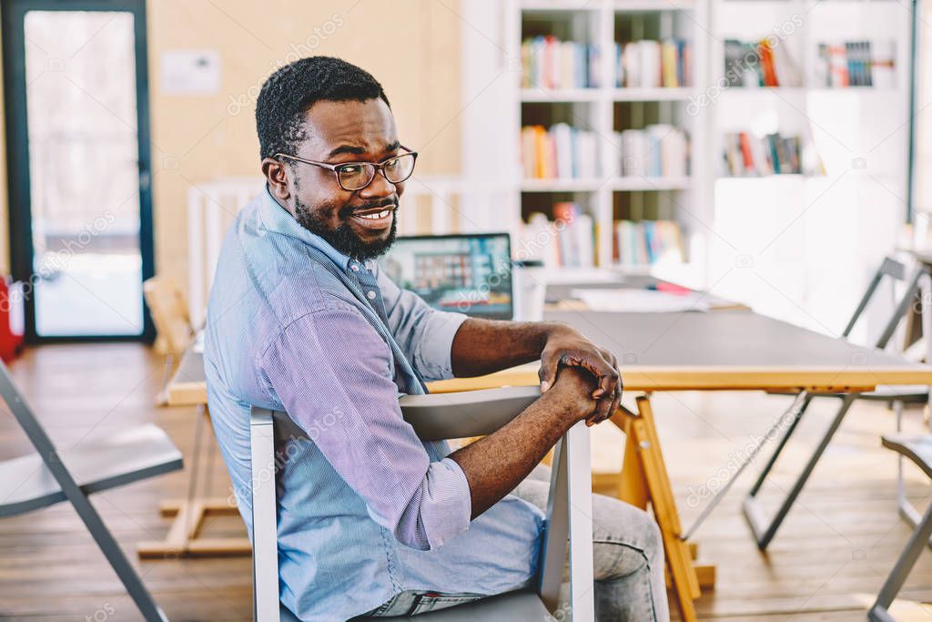Portrait of cheerful african american male freelancer sitting at coworking space satisfied with job,prosperous dark skinned hipster guy in spectacles looking at camera spending time in offic